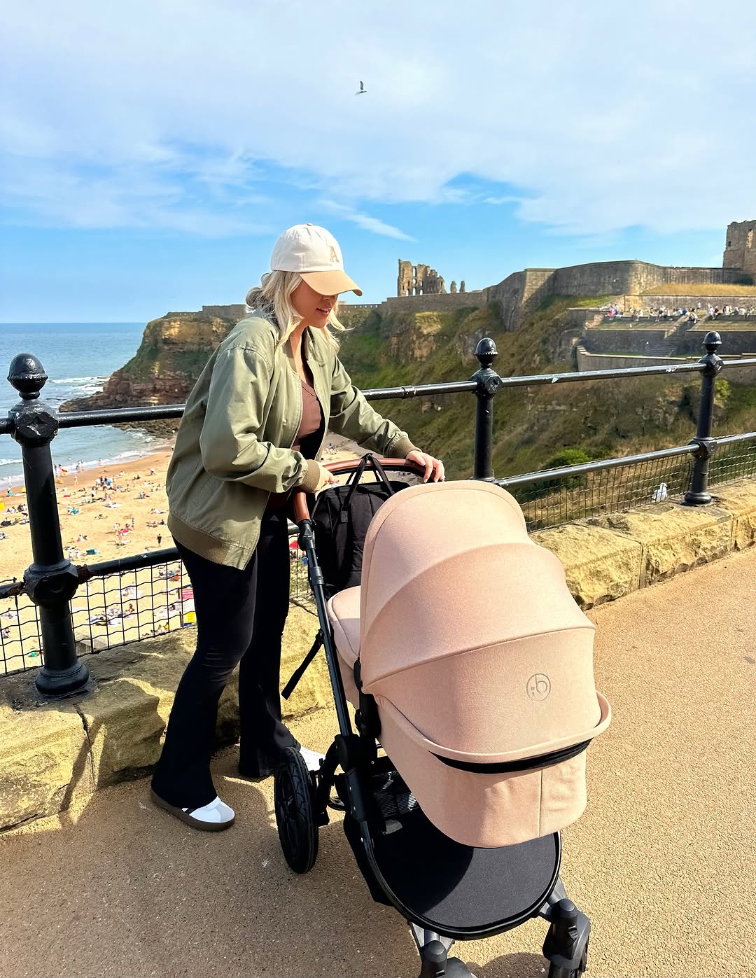 A woman in a beach town pushing a stroller along a coastal path. The Ickle Bubba stroller is a dusty blush pink with a black frame, and the background features a scenic view of cliffs, the sea, and a beach under a blue sky.