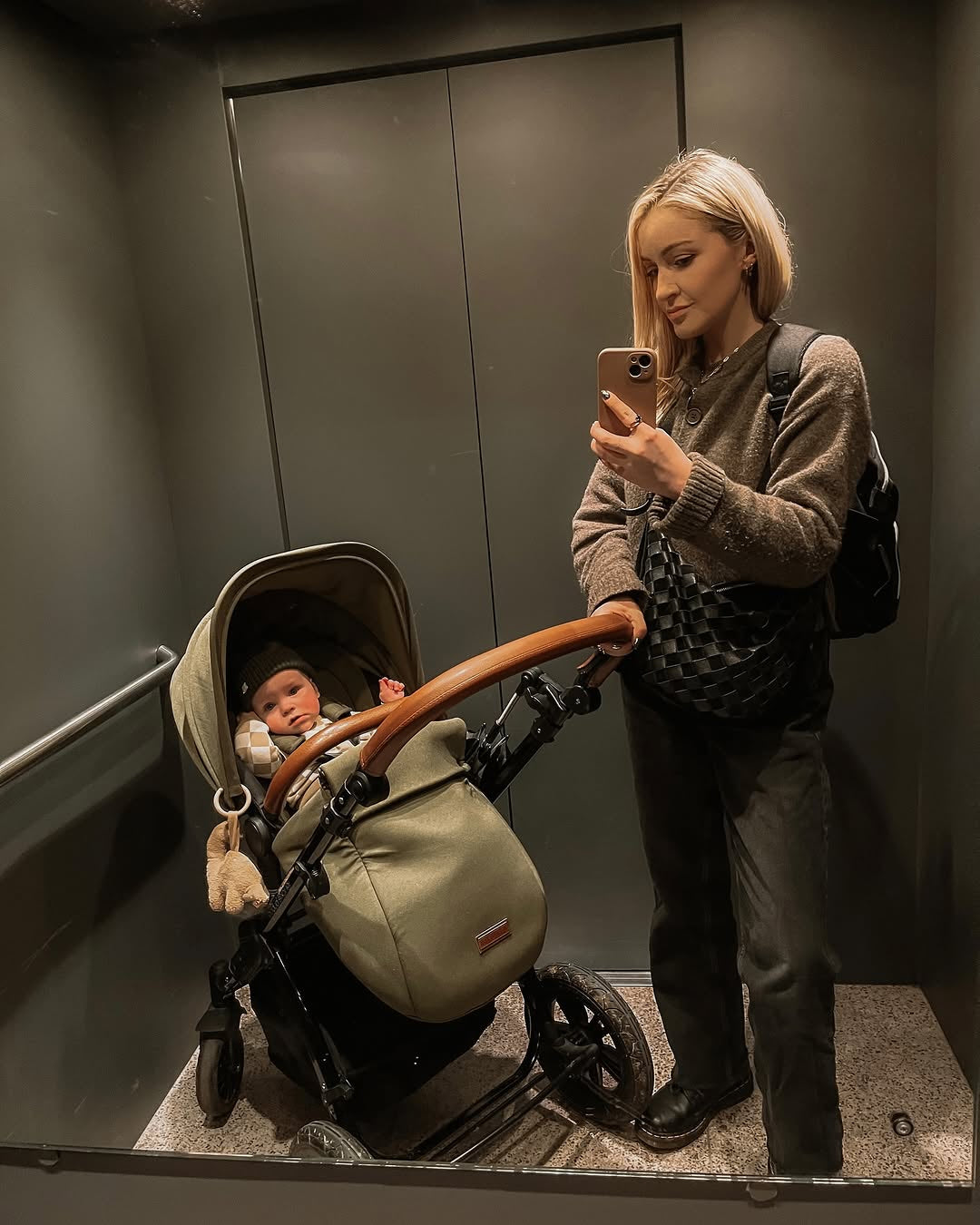 A woman takes a mirror selfie in an elevator with a baby in an Ickle Bubba stroller. The stroller is a sage green and black model with a brown wooden handlebar, and the baby is wearing a matching beige knit outfit.