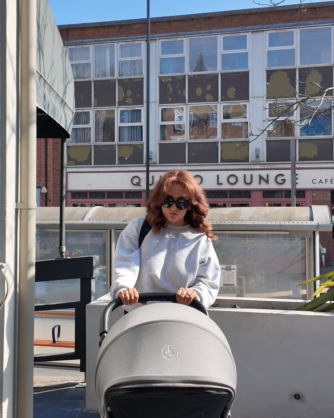 A woman with red hair wearing a light gray sweatshirt and sunglasses pushes a gray Ickle Bubba stroller outdoors in front of a building with a 'Quinto Lounge' sign.